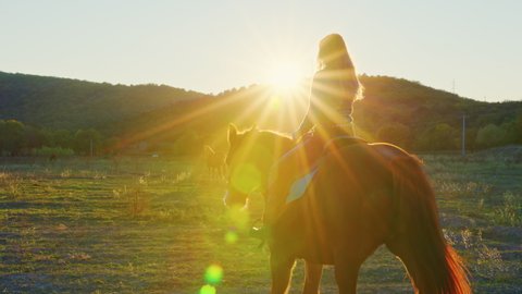 A woman rides a horse at sunset. A woman astride the horse slowly moves across the field with hills. Sun rays and glares