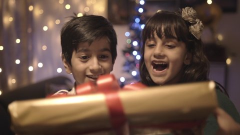 Two well dressed adorable young kids are smiling and receiving their Christmas present in a decorated house. Happy and elated cute girl and boy accepting the gift box from parents 