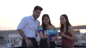 Group of three Indian friends having a video chat on a smartphone. Group of male and female friends waving hands to camera and happily talking on video call at rooftop cafe outdoors. 