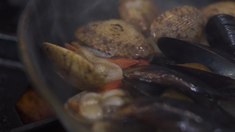 Preparing tasty mussels and scallops on metal frying pan in burning fire on contemporary stove in luxury seafood restaurant kitchen extreme close view.