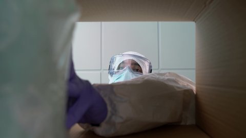 A male nurse in a protective suit, goggles and a mask is putting packets of masks into a cardboard box. Person preparing a package with medical supplies to combat Covid 19. View from inside package.