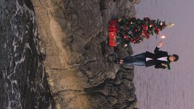 Young beautiful female with sparkler celebrating new year or Christmas at sea beach. Decorated Christmas tree at rock beach near sea. Concept of celebrating Christmas on a distance. Vertical video