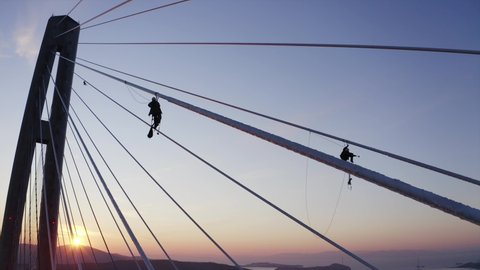 Aerial view of workers silhouettes hanging in safety harnessing on the Russian bridge cables cleaning them from ice after icy storm. Sunset. Vladivostok