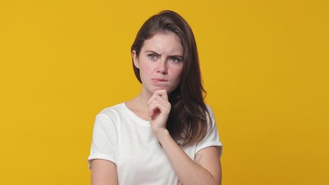 Puzzled pensive brunette young woman 20s years old wearing basic white t-shirt posing isolated on yellow color background in studio. People lifestyle concept. Looking aside up put hand prop up on chin