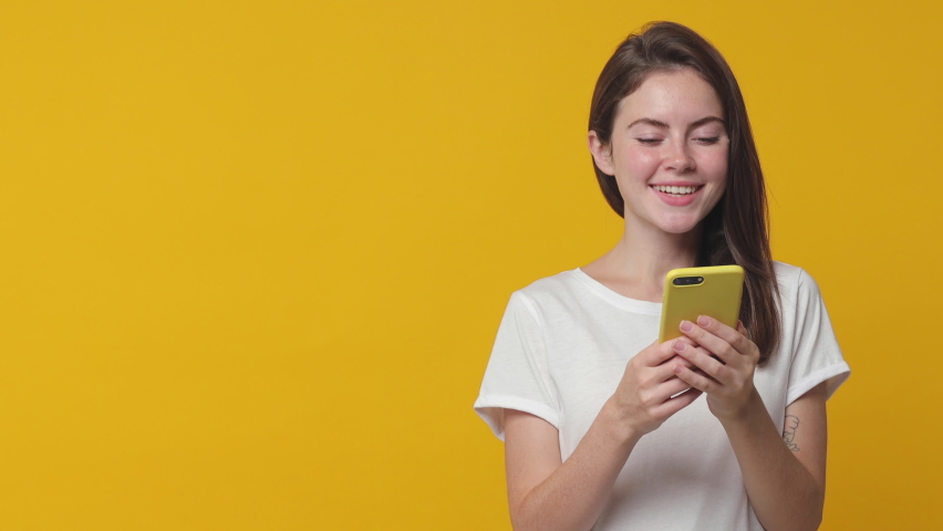 Excited brunette young woman 20s in white t-shirt isolated on yellow background in studio. People lifestyle concept. Point index finger on mobile cell phone aside typing sms message showing thumb up Royalty-Free Stock Footage #1063388653