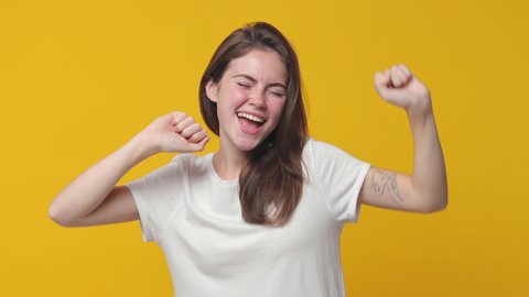 Cheerful funny brunette young woman 20s years old in casual white t-shirt posing isolated on yellow color background in studio. People lifestyle concept. Dancing clenching fists rising waving hands