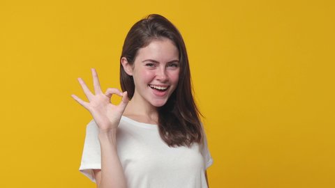 Excited cheerful brunette young woman 20s years old wearing basic white t-shirt posing isolated on yellow color background studio. People lifestyle concept. Looking camera showing OK gesture blinking