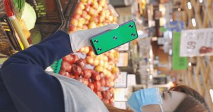 vetical video - close up of asian woman holds smart phone with green screen and shopping cart in fruit aisle at grocery store