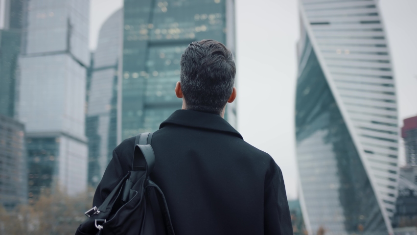 Man in coat looking at the skyscrapers pan shot right to left. Gimbal back shot of businessman in eyeglasses near skyscrapers in business city | Shutterstock HD Video #1063393882