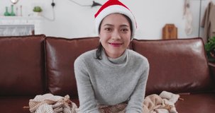 one lovely happy asian woman sitting in sofa wear red Christmas hat talking to webcam at home Lovely lady chatting online to friends say Merry Christmas prepare gift bag