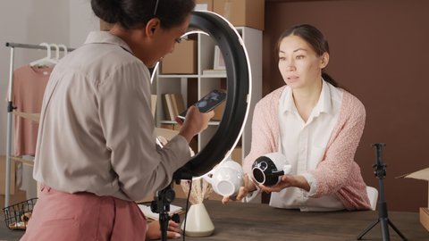Tracking shot of cheerful black woman standing by ring light and taking photos of cups held by her Asian female friend
