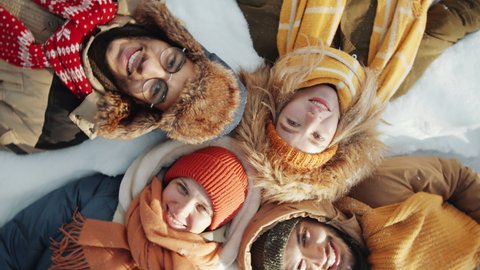 Top down shot of young cheerful multiethnic friends in outerwear lying together on snow, looking at camera and smiling while posing outdoors on winter day