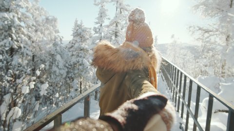 Point of view shot of young pretty woman holding hand of her boyfriend, smiling and talking to him while walking together in park on snowy winter day