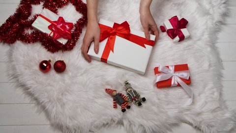 Womens hands place a gift box on a white fur skin, around a scattered red tinsel and decorations for the Christmas tree. Winter Holidays Christmas and New Year. Close up. Slow motion.