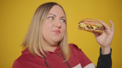 The plump woman points her eyes at the burger and takes a savory bite of it. Body positive. High-calorie food. Fast food