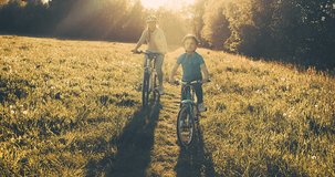 Mother teaching son to ride bicycle. Happy cute boy in helmet learn to riding a bike in park on green meadow in summer day at sunset time. Family weekend. 4K video Slow motion