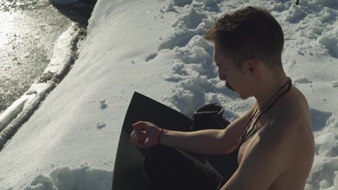 Man sitting on black yoga mat in snow with no shirt breathing heavily to prepare mind and body for frigid plunge into frozen mountain lake. Sun on shirtless guy hyperventilating before extreme sport.