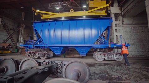 A worker at a modern train factory controls crane putting blue wagon on a wheels. Vehicle production. Advanced railroad car manufacturing plant. Tracking shot. Slow motion