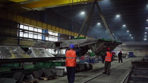 Two factory workers load with crane and control the huge metal construction of future train wagon. Modern workshop for the production of railway cars. Rail Car manufacturing Plant. Wide shot.