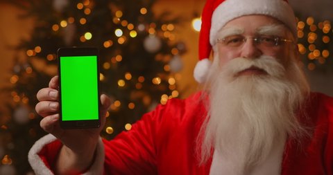 Santa Claus is sitting on the sofa in the background of a Christmas tree and garlands holding a mobile phone with a green screen pointing at it with his finger.