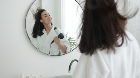 Slow motion of happy young woman drying hair using electric blow dryer and dancing in bathroom having fun. Emotions and modern lifestyle concept.