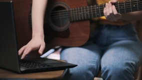 Online learning to play musical instruments. A woman is typing on a laptop, holding a guitar. Close up of hands.