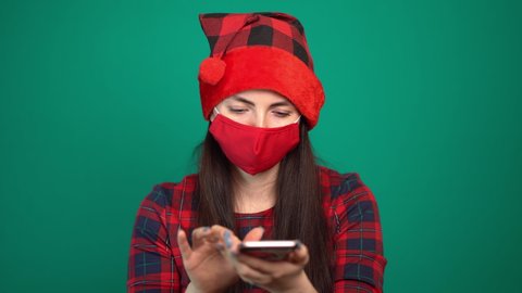 woman in santa claus hat, wearing a medical mask on her face, writes a message on a smartphone or happy new year.