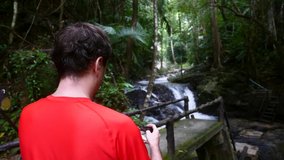 Travel Man in Red T-shirt Using Mobile Phone and Shooting Video with Waterfall and Beautiful Nature in Green Tropical Forest Public Park in Thailand. Tourist Make Video Content for His Blog