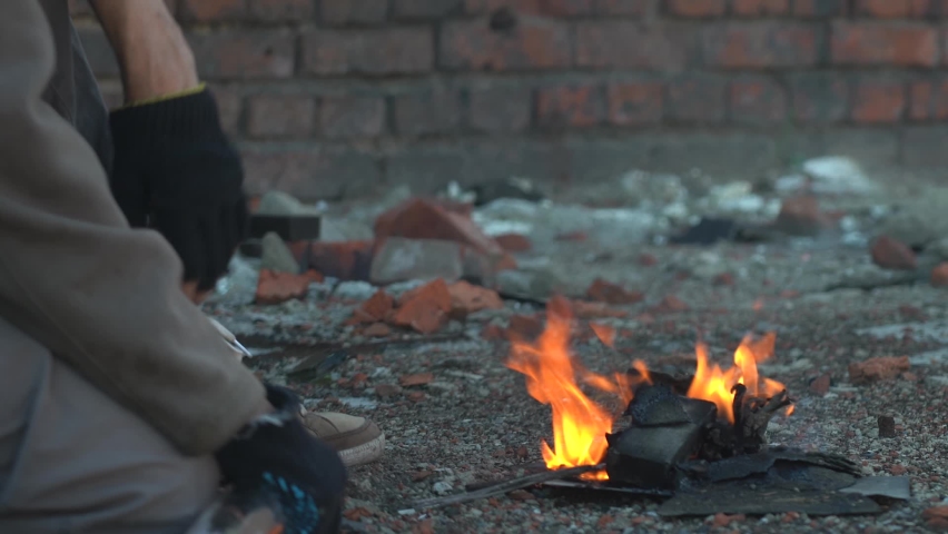 A homeless man warms his hands by the fire. Post-apocalypse scene. Hands in black torn gloves. Close-up. Weak warmth from a small fire. Royalty-Free Stock Footage #1063422994