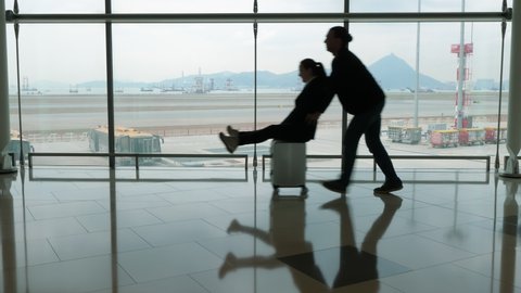 Funny couple at airport, woman race on trolley case, man run behind and push rider forward. Silhouetted shot against terminal window. Passengers have fun after long flight arrival, enjoy like kids