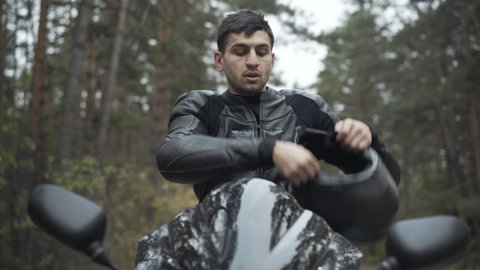 Portrait of handsome Middle Eastern man putting on motorcycle helmet and leather biker gloves. Confident serious male motorcyclist sitting on motorbike. Racing and lifestyle concept.