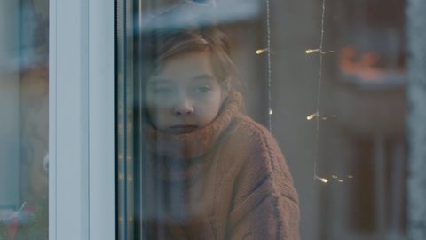 Serious sad little teenager girl in sweater standing behind window waiting for winter holidays christmas staying home alone. Childhood. December.