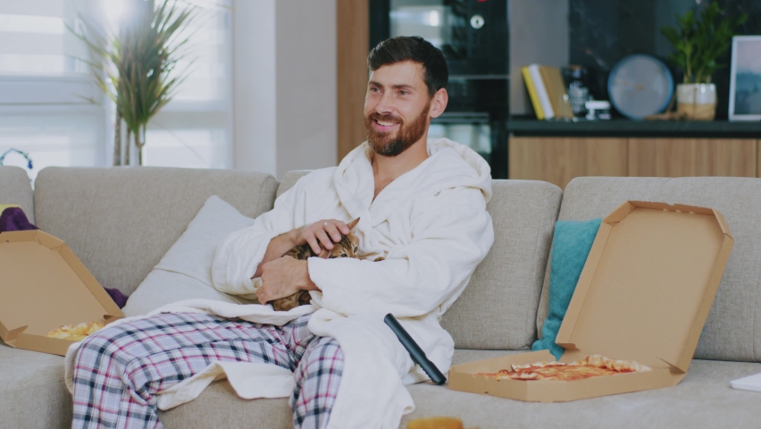 Happy young man with cat watching TV eating pizza. Sitting on sofa at home. Relaxation cinema channels resting. Portrait. Close up. Slow motion. Royalty-Free Stock Footage #1063427083