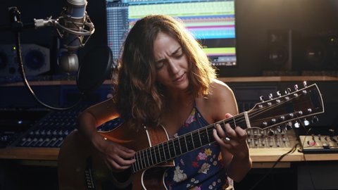 Music studio. Creative world. Musician. Latin woman playing guitar and singing in the recording studio. An attractive brunette female person in a beautiful shirt records songs. 