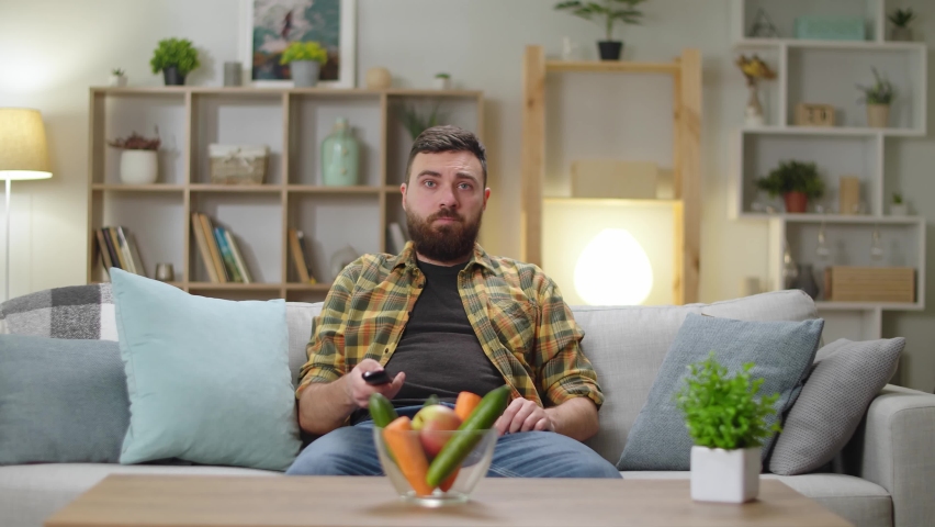 Young handsome man sitting on a couch at home and watching tv | Shutterstock HD Video #1063430818