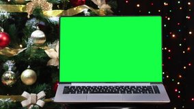 Modern laptop with green screen and Christmas tree with ornaments and blinking lights