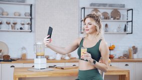 fitness young woman posing for photo with glass of fresh vegetable fruit juice in kitchen interior. Happy woman drinking healthy drink in front of camera phone. Pretty girl making video on smartphone