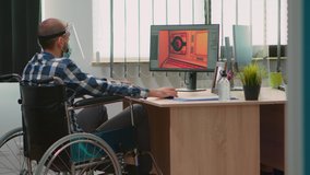 Handicapped game developer sitting in wheelchair with protection mask working at new project from new normal studio office during covid-19 pandemic. Immobilized man respecting social distance.