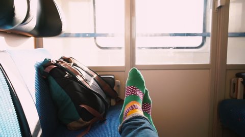 The traveler's feet in socks lie on seats in the train. Nearby is backpack. Light movement in the window. Carriage passengers in public transport. View from the eyes. Relaxation while traveling.