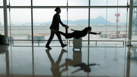 Travelers couple have fun, woman ride lying on trolley case. Man walk behind, hold legs and push forward. Silhouetted shot against glass wall at terminal room