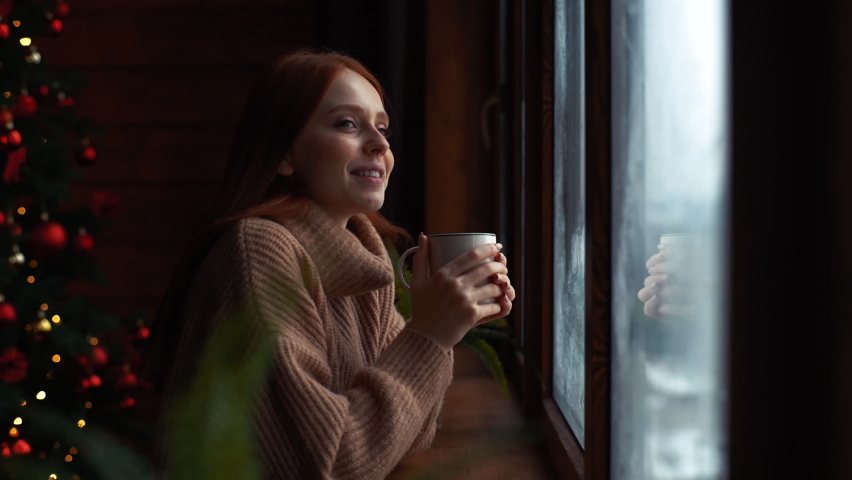 Side view of happy young redhead woman with cup standing by window at home at Christmas time. Beautiful charming lady drinking coffee on xmas Eve at cozy living room with festive interior. Royalty-Free Stock Footage #1063446028