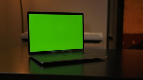 Notebook showing green screen stands on a desk in living room, copy space for pasting. Cozy living room in the evening with warm lights In the background.