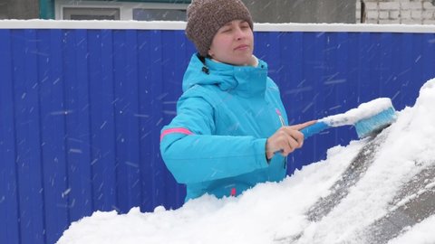girl sweeps snow from a car glass. A woman cleans the car of snow with a brush. Scraping snow and ice from a car windshield. Snowfall covered the car.