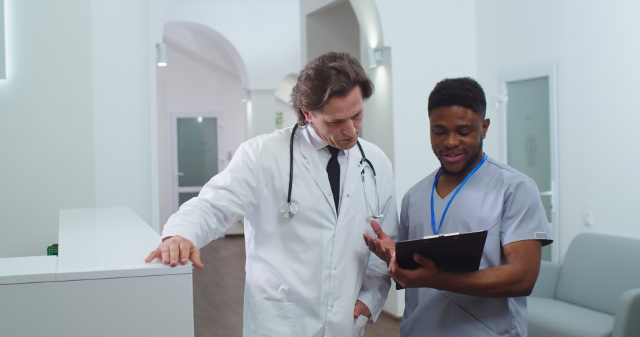Portrait of African American male nurse standing in hospital and speaking with Caucasian doctor about treatment results. Healthcare professionals talking in clinic. Medical concept | Shutterstock HD Video #1063447360