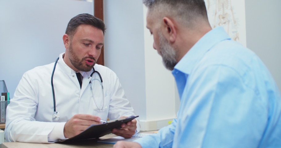 Portrait of male Caucasian doctor in white coat speaking with senior patient man while sitting in hospital cabinet. Rear of male taking to physician at consultation. Doctor appointment concept | Shutterstock HD Video #1063447546