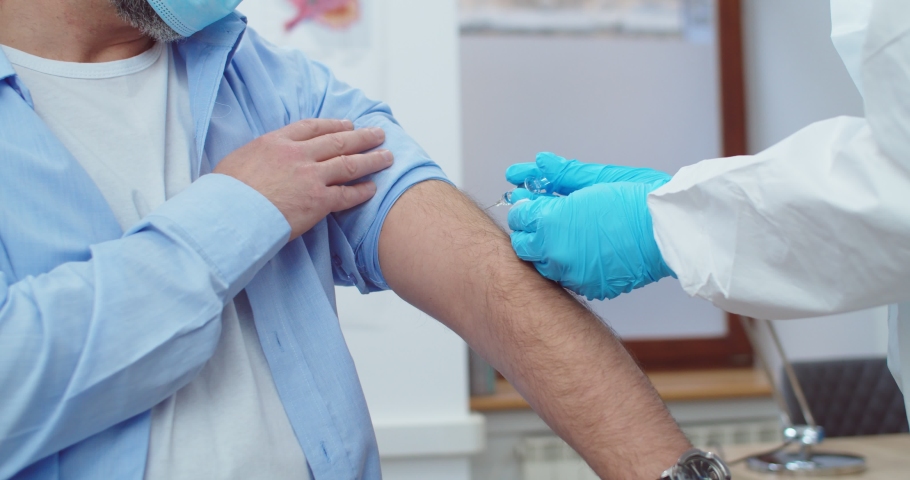 Close up shot. Professional doctor hands in sterile protective uniform and gloves making injection to senior Caucasian patient man in hospital office. Coronavirus pandemic. Analysis concept | Shutterstock HD Video #1063447606