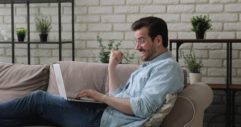 Emotional young man lying on cozy sofa, received online lottery win email notification on computer, celebrating personal success, getting dream job offer or banking credit mortgage loan approval.