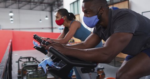 Diverse woman and man wearing face masks exercising at gym. working out on exercise bikes. hygiene at gym during coronavirus covid 19 pandemic : vidéo de stock