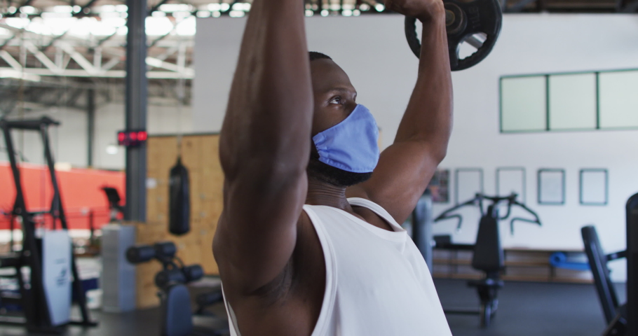 Fit african american man wearing face mask exercising using barbell in the gym. social distancing quarantine lockdown during coronavirus pandemic