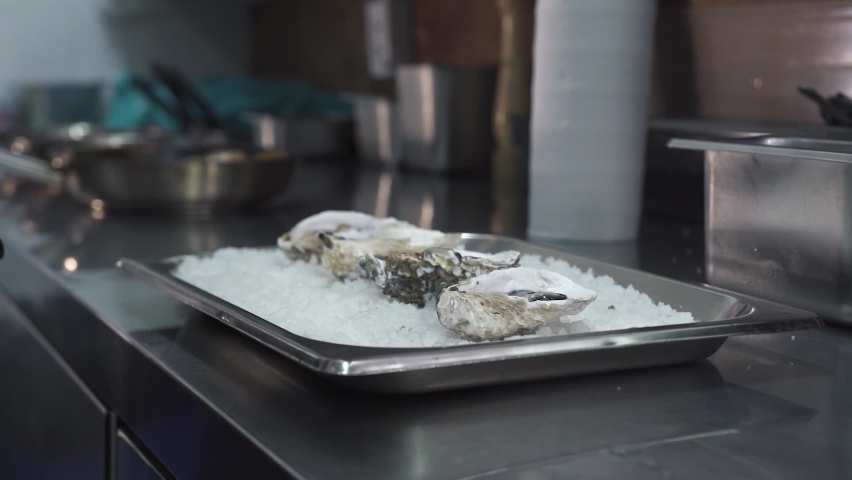 Man in gloves adds spicy sauce to delicacy oysters on metal tray with heap of crushed ice at countertop in kitchen of seafood restaurant close view slow motion.